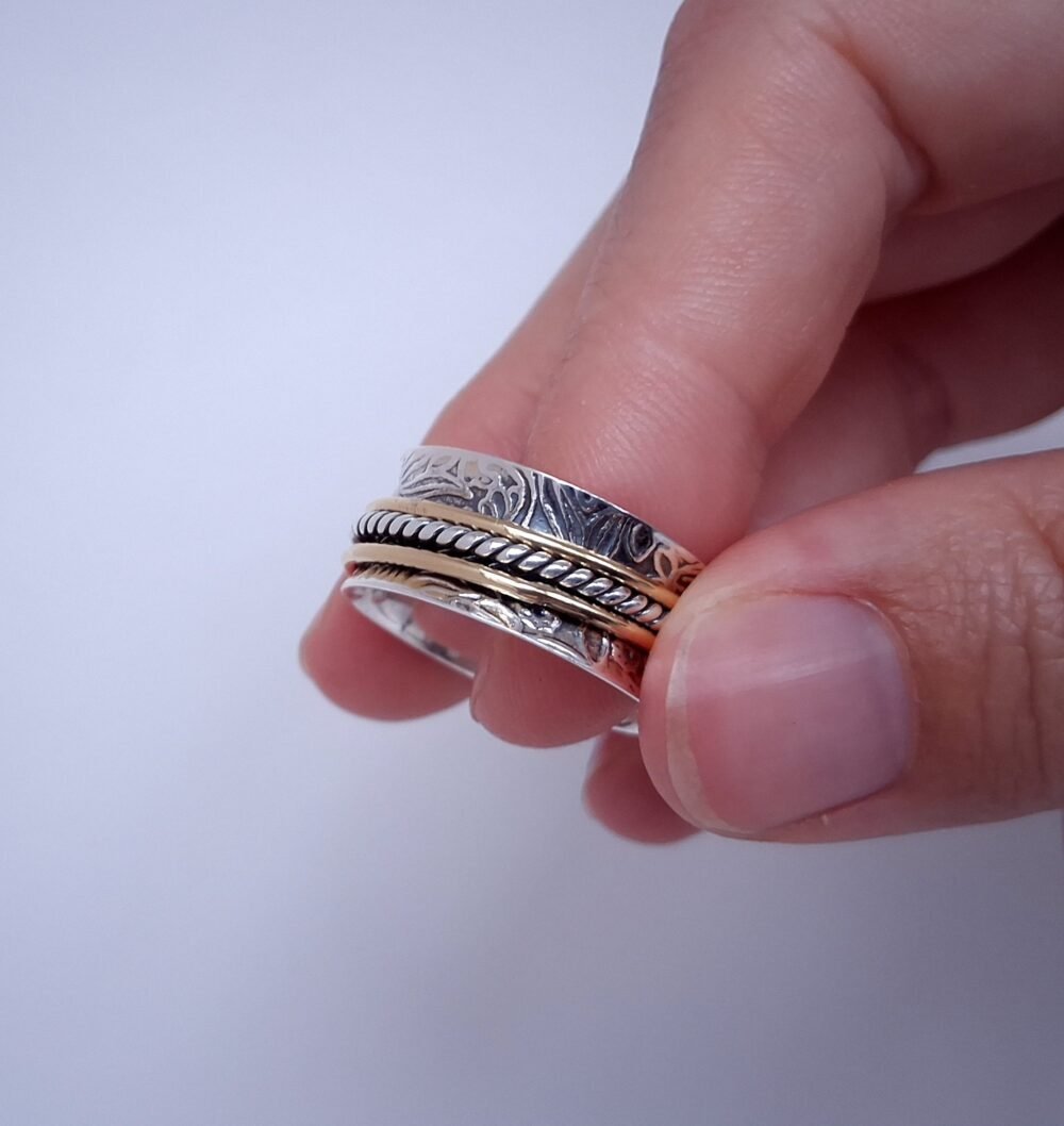 Detailaufnahme eines Drehringes aus Silber. Das Hauptband ist konkav und mit einem floralen Muster versehen. Darüber gleiten 3 dünne Ringe in Gold und Silber, die unabhängig voneinander gedreht werden können. Der Ring wird von einem Model an den Fingerspitzen gehalten. Weißer Hintergrund.
